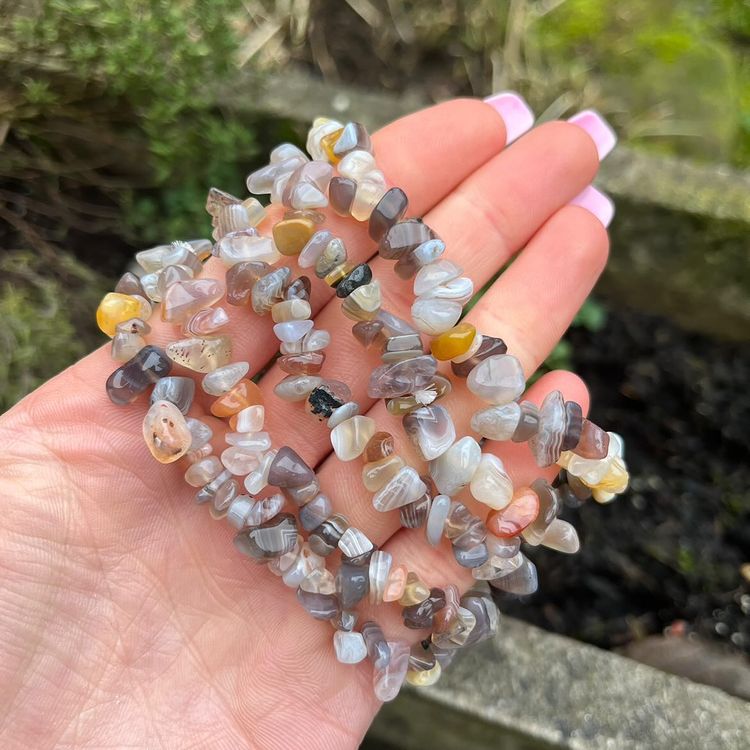 An open palm holding several Agate crystal chip bracelets outdoors, showcasing the polished, irregular gemstone beads. The stretchy bands feature unique earthy grey, soft cream, and muted mustard yellow colour bands, shot against a blurred garden backdrop to highlight a comforting and grounded everyday style.