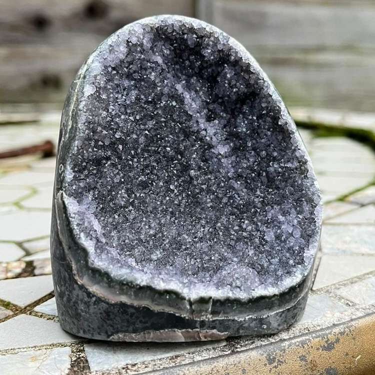 A crisp front view of a substantial Amethyst cut base point crystal geode dome standing upright outdoors on a stone mosaic patio table. The premium gemstone specimen features a flat cut base, a smoothly polished outer rind, and a hollow core packed with thousands of tiny, sparkling deep purple druzy crystal points to inspire an evening meditation ritual.