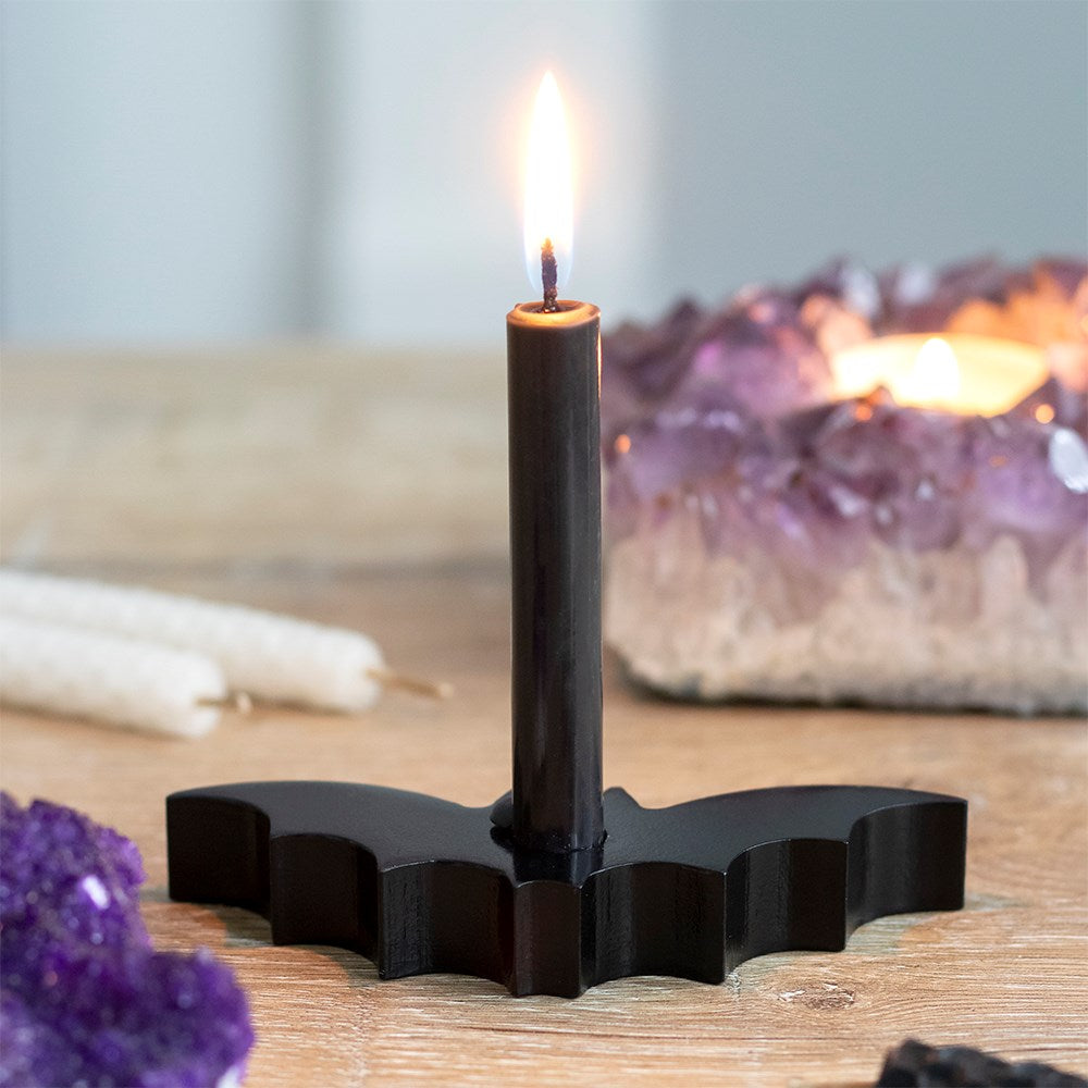 An atmospheric lifestyle shot of a glossy black resin bat spell candle holder resting on a rustic wooden tabletop surface. A tall, black taper candle burns brightly in the centre slot, styled beautifully in front of a massive raw purple Amethyst crystal cluster geode and unlit white chime candles.