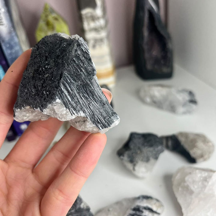 A close-up view of a hand holding a jagged specimen of black tourmaline matrix nestled inside a rough white quartz base, perfect for styling by a front door or on a busy office desk workspace layout.