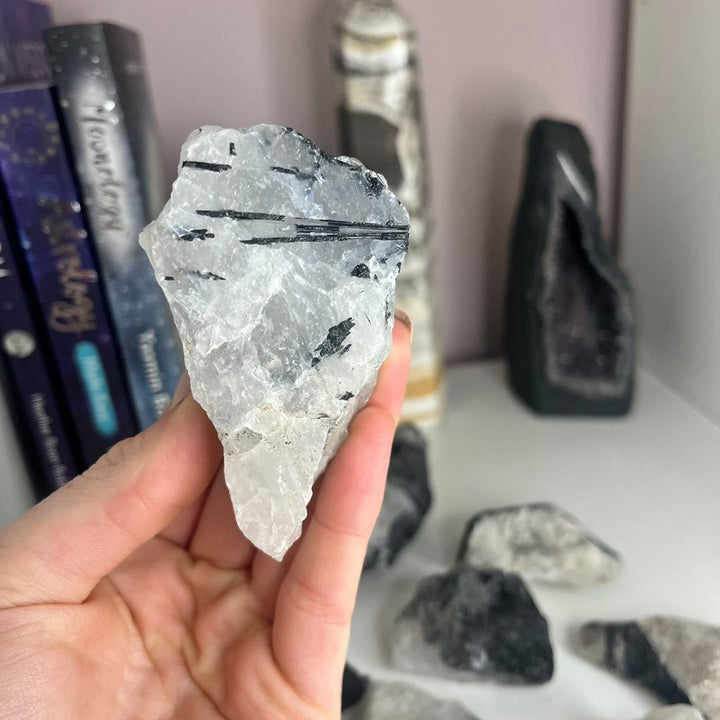 A hand displaying a raw tourmaline quartz point against a blurred background of towers, detailing fine, dark black tourmaline strands growing naturally inside an icy crystalline structure to assist with spiritual clarity.