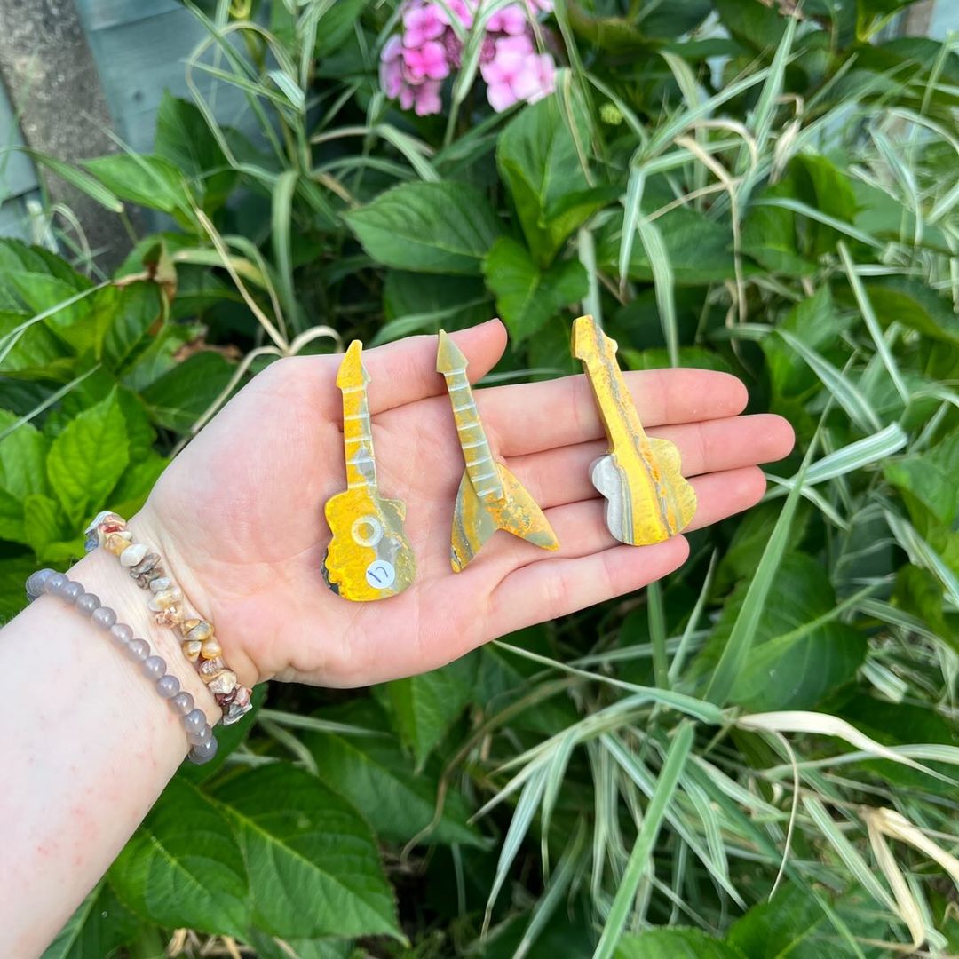 An open hand wearing layered crystal chip bracelets presenting three unique bumblebee jasper guitar crystal carvings outdoors, showcasing vibrant yellow bands, dark charcoal tiger patterns, and pale grey volcanic layers against a lush green garden backdrop for Solar Plexus Chakra alignment.