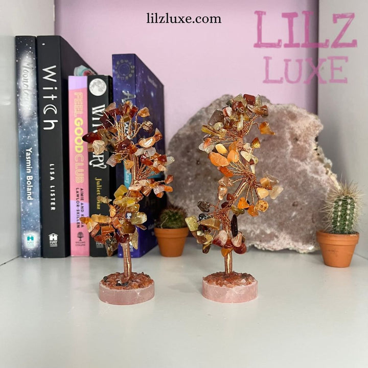 Two handmade Carnelian crystal trees standing on a white shelf, showcasing flexible wire branches laden with glossy red-orange gemstone leaves and a solid pink quartz base scattered with tiny raw mineral chips against a backdrop of books and small cacti.