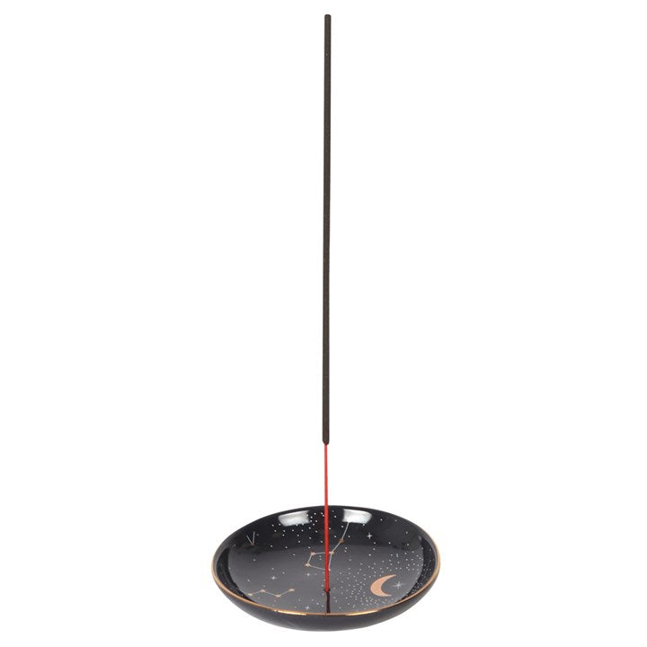 A straight-on studio shot showing a shallow black ceramic constellation incense burner tray with a single long brown incense stick inserted centrally. The minimalist graphic layout showcases the circular ash catcher plate against a clean white backdrop, highlighting its smooth glaze and cosmic design for evening relaxation.
