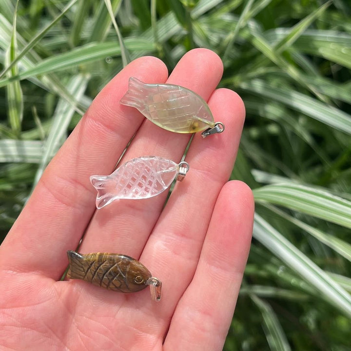 A person holding miniature polished crystal animal charms in their hand, including a translucent Clear Quartz fish pendant.