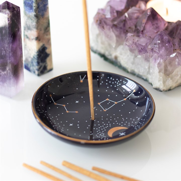 A lifestyle close-up view of a round, glossy dark blue ceramic constellation incense stick holder with an upright burning incense stick. The dish features a gold foil night sky pattern with a crescent moon and star signs, arranged on a white surface alongside a large raw purple Amethyst druzy cluster and polished crystal towers to invite a celestial calm.
