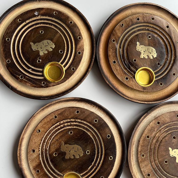 An overhead shot of four round mango wood incense holders designed for sticks and cones, showcasing concentric ring grooves, multiple pre-drilled stick guide holes, a small brass cup for burning incense cones safely, and a detailed elephant carving in the centre.