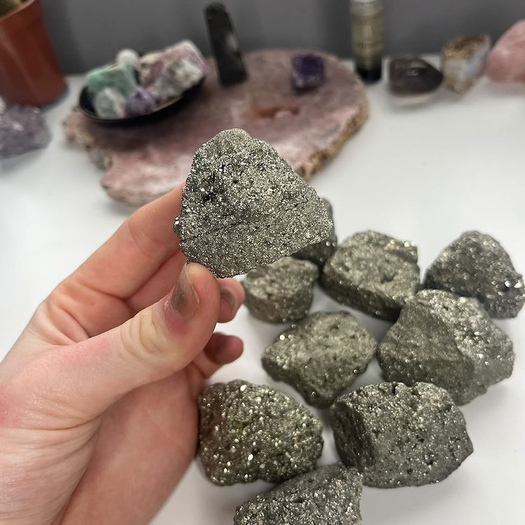 A hand lifting a single glittering raw pyrite crystal specimen from a desktop display collection of metallic gold chunks.