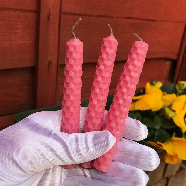 A close-up view of three pink honeycomb beeswax spell candles held upright in a white gloved hand outdoors. The hand holds the rolled textured candles firmly against a background of an outdoor reddish-brown wooden fence and yellow garden flowers to welcome cosy intentions and Heart chakra harmony.