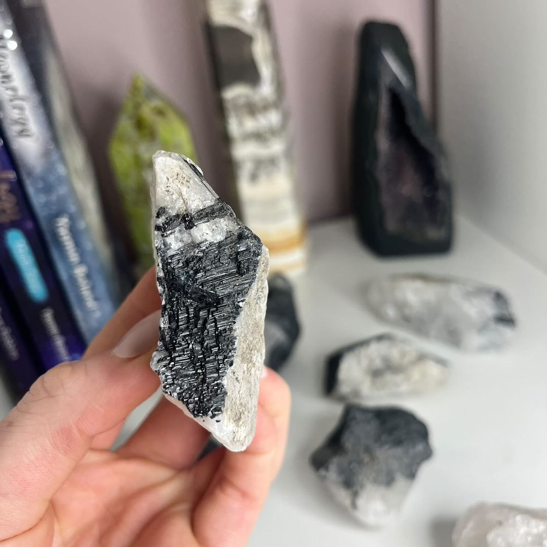 A hand showing a vertical view of a geological tourmalinated quartz stone, illustrating the distinct contrasting layers of glass-like quartz and dark earthy mineral growth points used to naturally cleanse a room&
