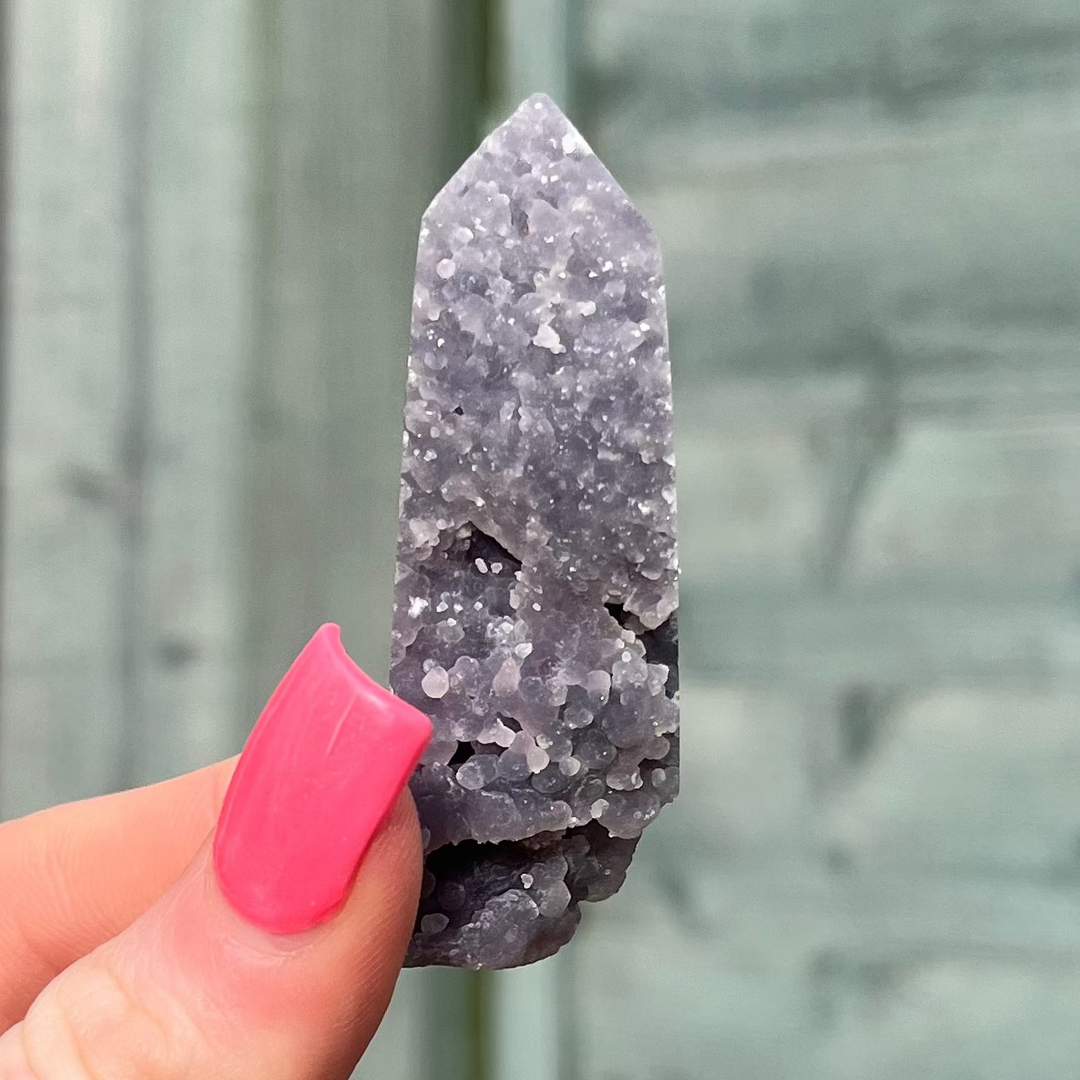 A close-up view of a hand with pink nails holding a polished grape agate crystal tower point, showcasing its velvety purple hue, intricate botryoidal clusters, and matte texture against a soft grey garden fence background for Crown Chakra healing.