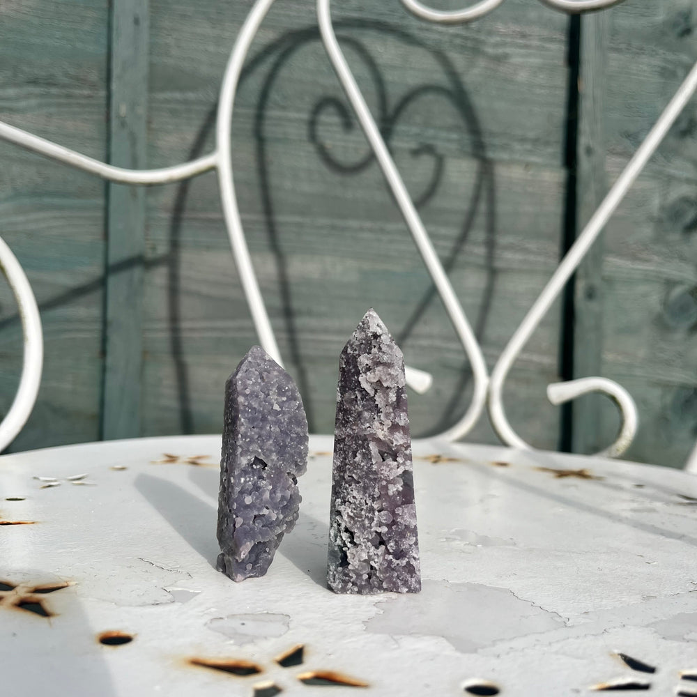 Two majestically sculpted grape agate towers standing upright on a decorative white metal garden table, highlighting the natural grape-like formations and unique colour zones that bring a sustaining aura of deep emotional peace to a ritual space.