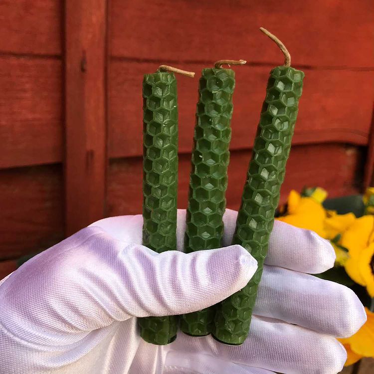 A close-up view of three green honeycomb beeswax spell candles held vertically in a white gloved hand. The hand displays the textured, hand-rolled candles against a backdrop of outdoor wooden panelling and yellow garden flowers to welcome fresh growth and Heart Chakra abundance.