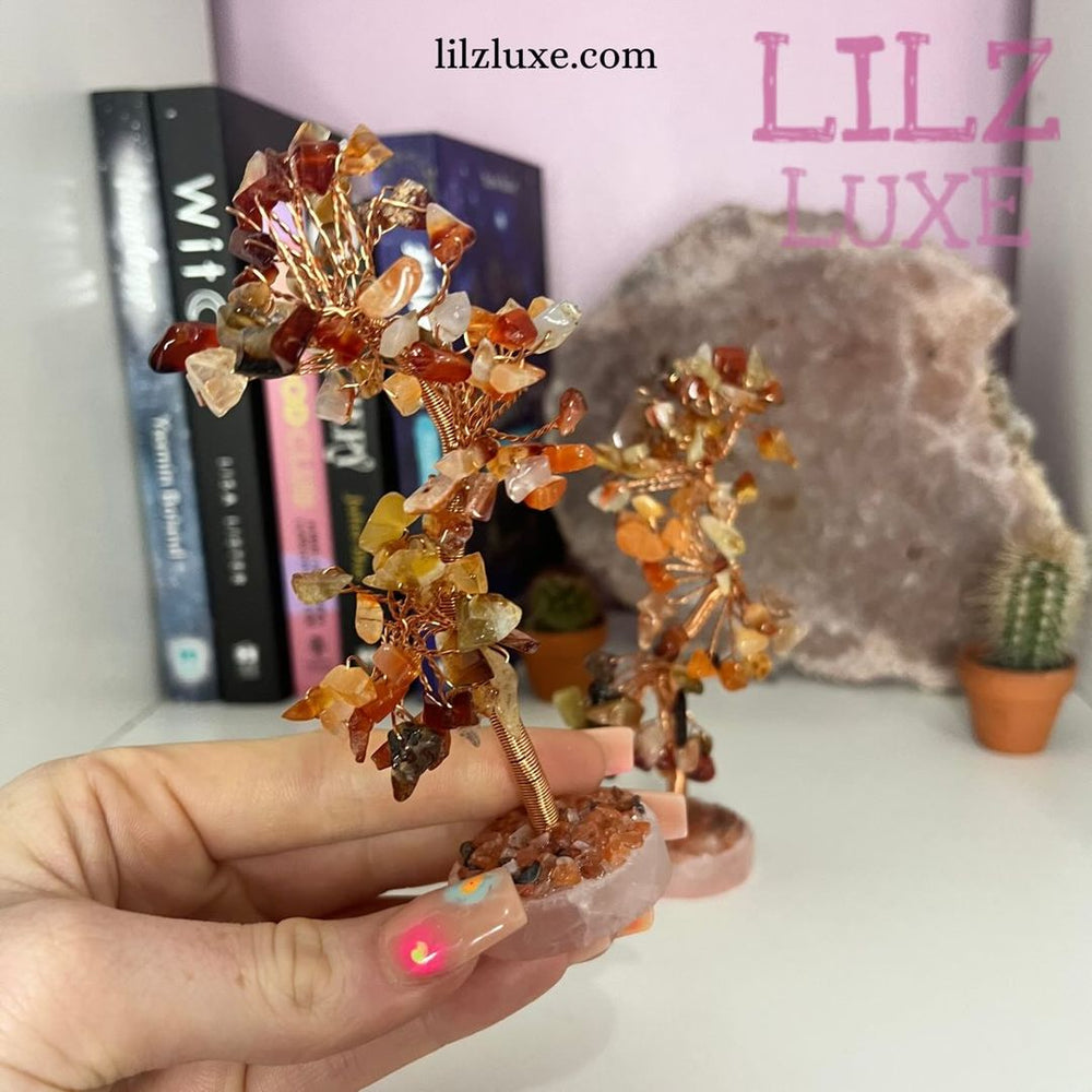 A hand holding a compact 10-centimetre Carnelian crystal wire tree indoors, displaying its custom-shaped branches filled with rich orange and red tumbled stone chips to illustrate its small shelf-decor scale.