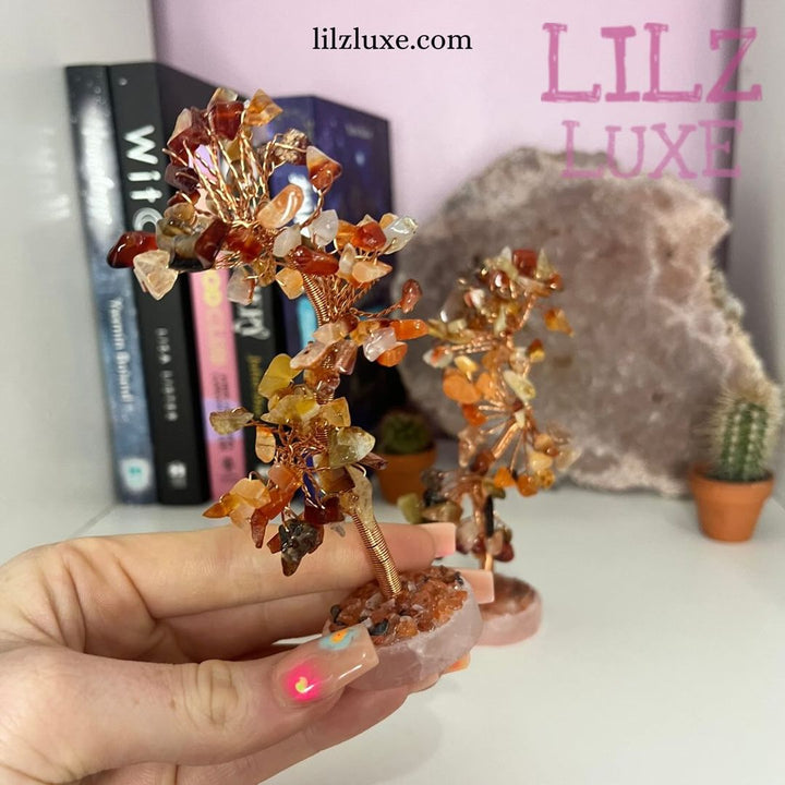 A hand holding a compact 10-centimetre Carnelian crystal wire tree indoors, displaying its custom-shaped branches filled with rich orange and red tumbled stone chips to illustrate its small shelf-decor scale.