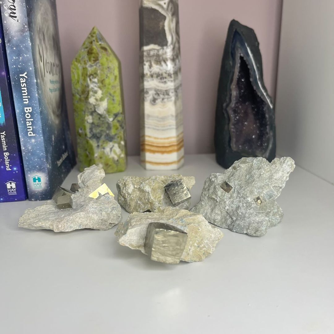 A group display of raw imperfect pyrite in matrix crystal specimens arranged on a white shelf, showcasing gritty, textured host rock blocks filled with glittering cubes of metallic gold pyrite in front of spiritual books and crystal towers for Solar Plexus Chakra alignment.