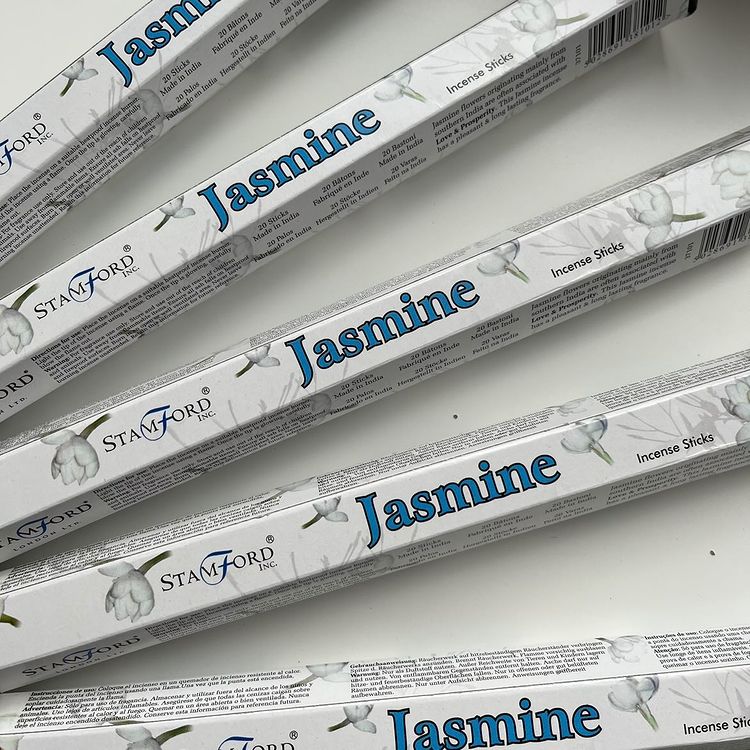 A close-up, top-down view of multiple hexagonal boxes of Stamford Jasmine incense sticks neatly stacked together on a clean, minimalist off-white surface. The sleek packaging features detailed illustrations of delicate white jasmine flowers, highlighting the clean-burning floral quality crafted to release a rich, sweet aroma for a comforting Heart chakra meditation routine.