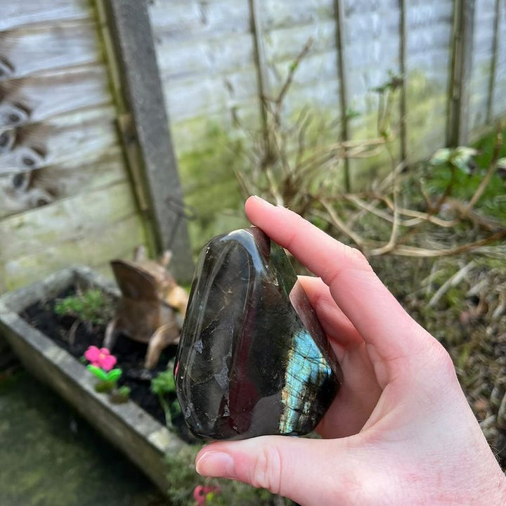 A side-angle profile shot of a polished labradorite freeform mineral specimen held outdoors, revealing dark grey crystalline structures and natural subterranean fracture lines.
