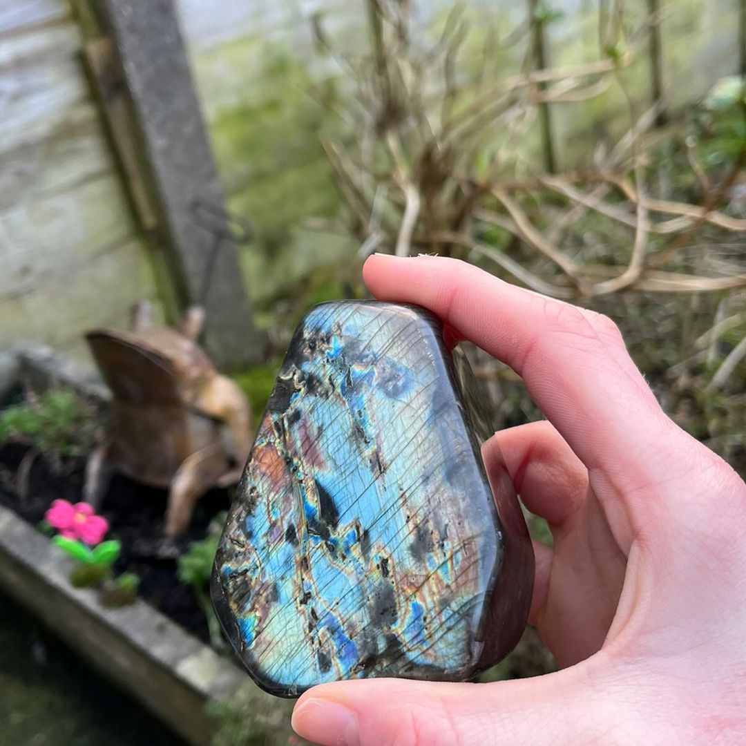 A hand steadying a self-standing polished labradorite freeform stone upright, highlighting its thick base and vibrant blue flashes in natural outdoor lighting.
