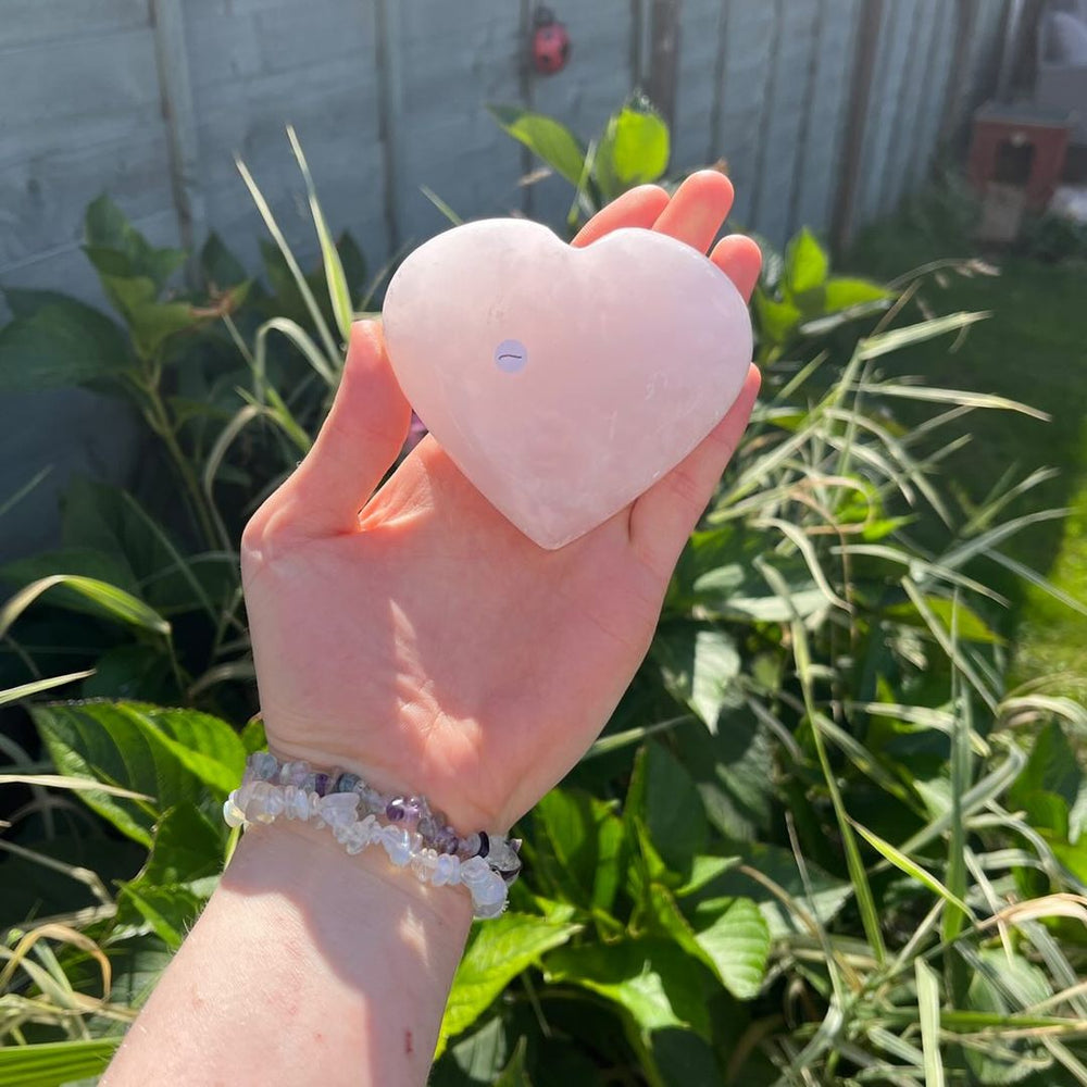 A hand presenting a single hand-carved pink mangano calcite heart outdoors under direct sunlight, illustrating its substantial 245-gram scale and smooth porcelain finish that brings a nurturing presence and peace into your home.
