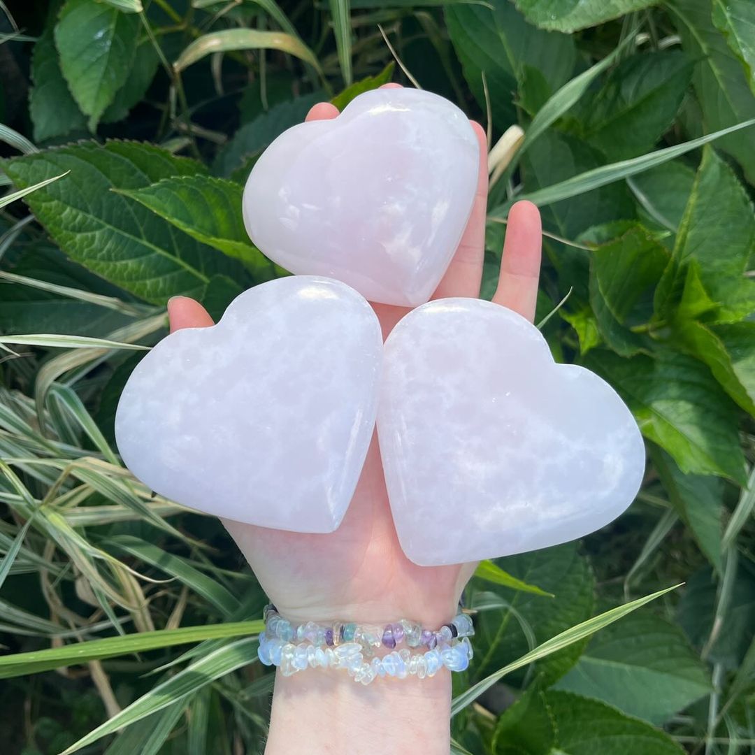 An open hand with a crystal chip bracelet cradling three polished mangano calcite hand-carved hearts outdoors, displaying their soft milky-pink shades and gentle organic lines against a backdrop of green garden leaves for Heart Chakra alignment.