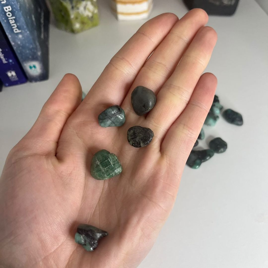 An open hand holding five polished mini emerald tumble stones over a white table setup, showcasing their compact 1cm sizing scale, rich mossy green hues, and smooth glossy textures for Heart Chakra alignment.
