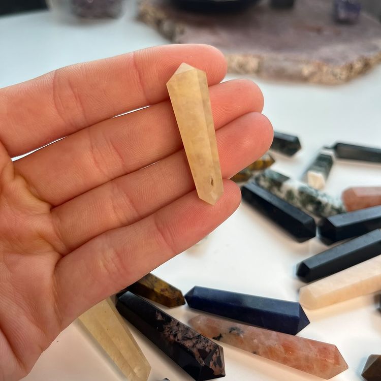 A hand holding a polished mini golden healer double-terminated point crystal close up, showcasing its translucent honey-yellow coloration and inner veils, styled indoors over a scattered layout of multi-coloured gemstone wands for Solar Plexus Chakra alignment.
