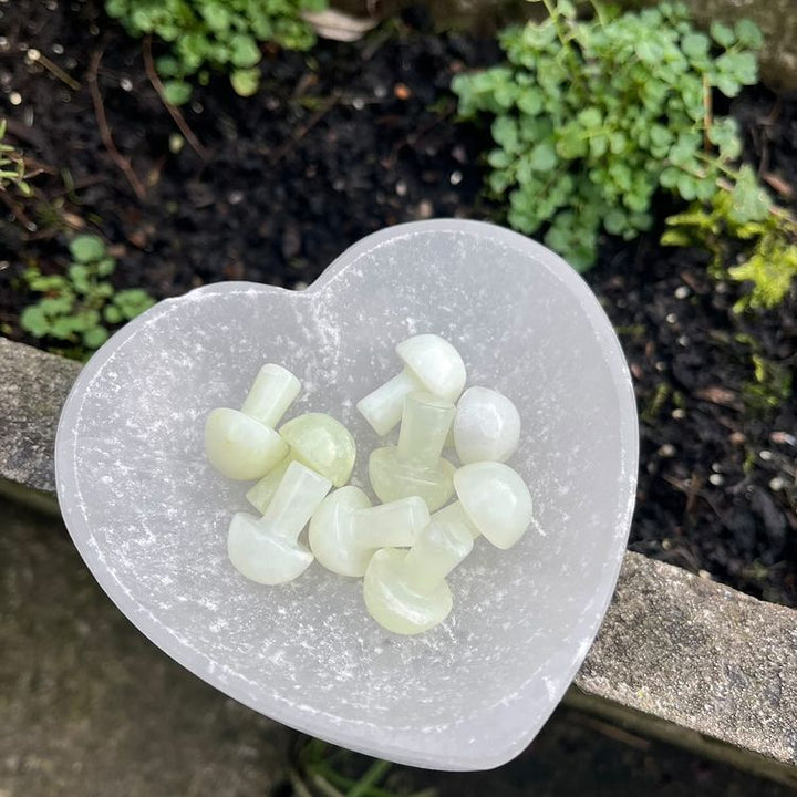 A collection of small, hand-carved light green Jade crystal mini mushrooms gathered inside a white satin spar selenite heart-shaped crystal bowl outdoors. The polished pieces showcase unique milky green swirls and natural patterns, photographed on a stone ledge next to fresh garden plants to promote an atmosphere of good luck and Heart chakra harmony.