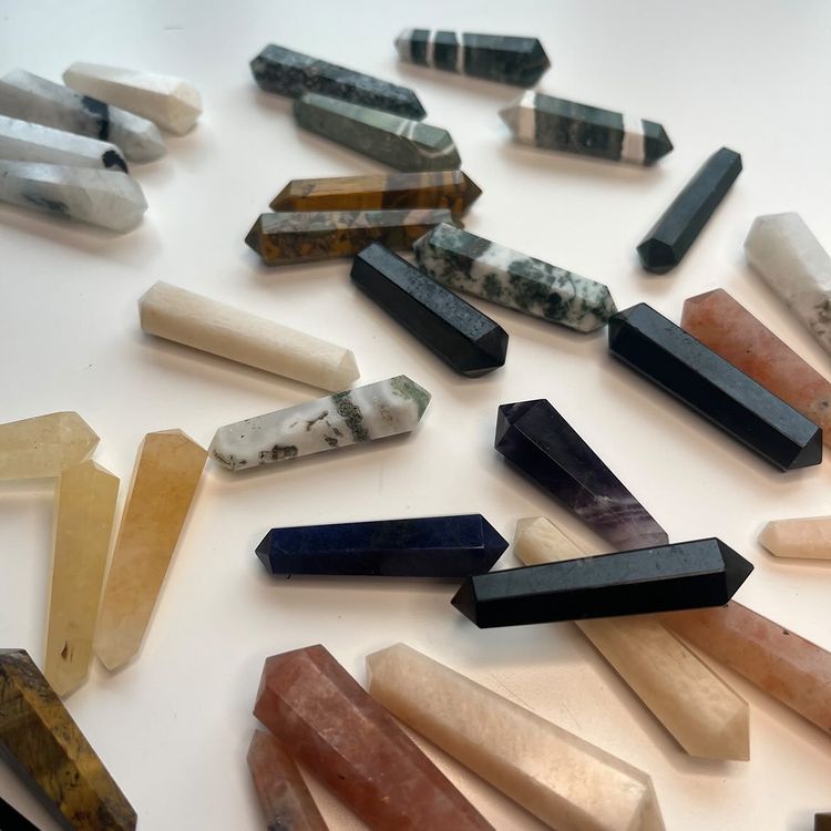 A scattered collection of polished mini mystery double terminated crystal points displayed on a white surface, showcasing a variety of minerals including tiger's eye, obsidian, amethyst, rose quartz, and sodalite, highlighting their dual-pointed 4cm shapes.