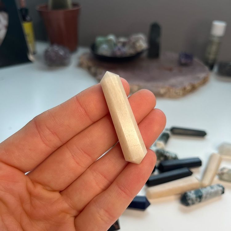 A hand holding a polished mini pink moonstone double-terminated point crystal close up, showcasing its dreamy pearlescent layers, soft blush-pink patches, and milky veins, styled indoors over a scattered layout of multi-coloured gemstone wands for Third Eye and Crown Chakra alignment.