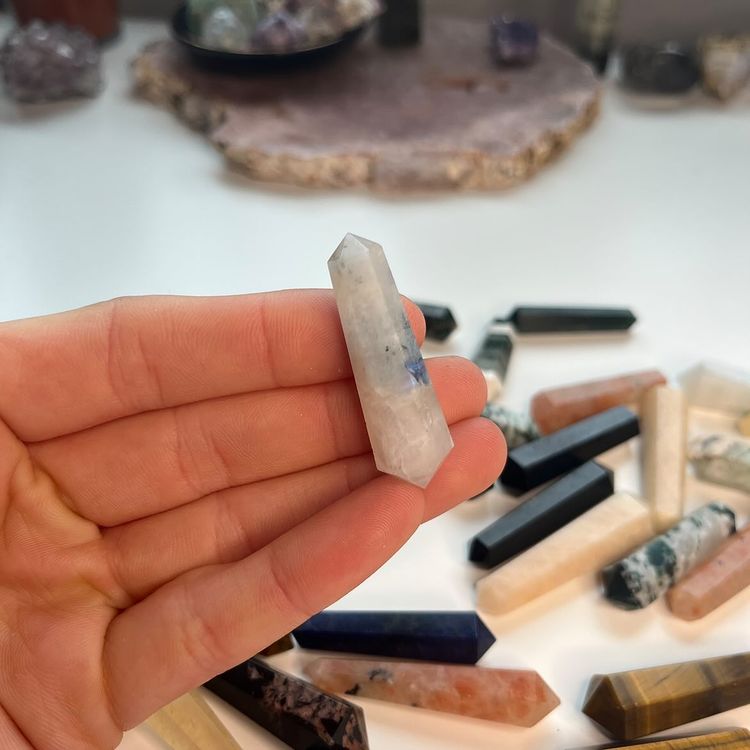 A hand holding a polished mini rainbow moonstone double-terminated point crystal, highlighting its milky white translucency and iridescent blue flash under ambient light, with a collection of multi-coloured gemstone points blurred on the desk background for Crown Chakra harmony.
