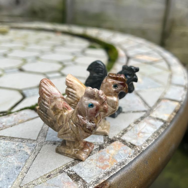 A close-up view of two intricately carved soapstone bird figurines standing in a row on a stone mosaic patio table outdoors. The foreground features a light tan and brown streaked cockerel, while a dark grey and black textured hen rests just behind it, showcasing the velvety smooth natural finish and organic colour variations that support Root chakra grounding.