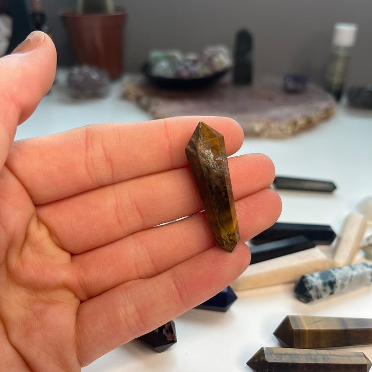 A hand cradling a polished mini tiger's eye double-terminated point crystal, showing off its rich silky texture and golden-brown bands of natural chatoyancy, styled indoors over a selection of multi-coloured gemstone wands for Solar Plexus Chakra alignment.