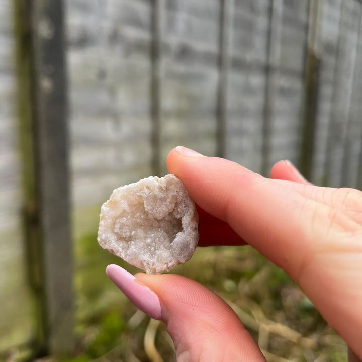 Fingertips gently holding a single open mini white quartz geode outdoors, highlighting its small pocket-sized scale, bumpy outer crust, and hollow glittering internal crystal cave under daylight.