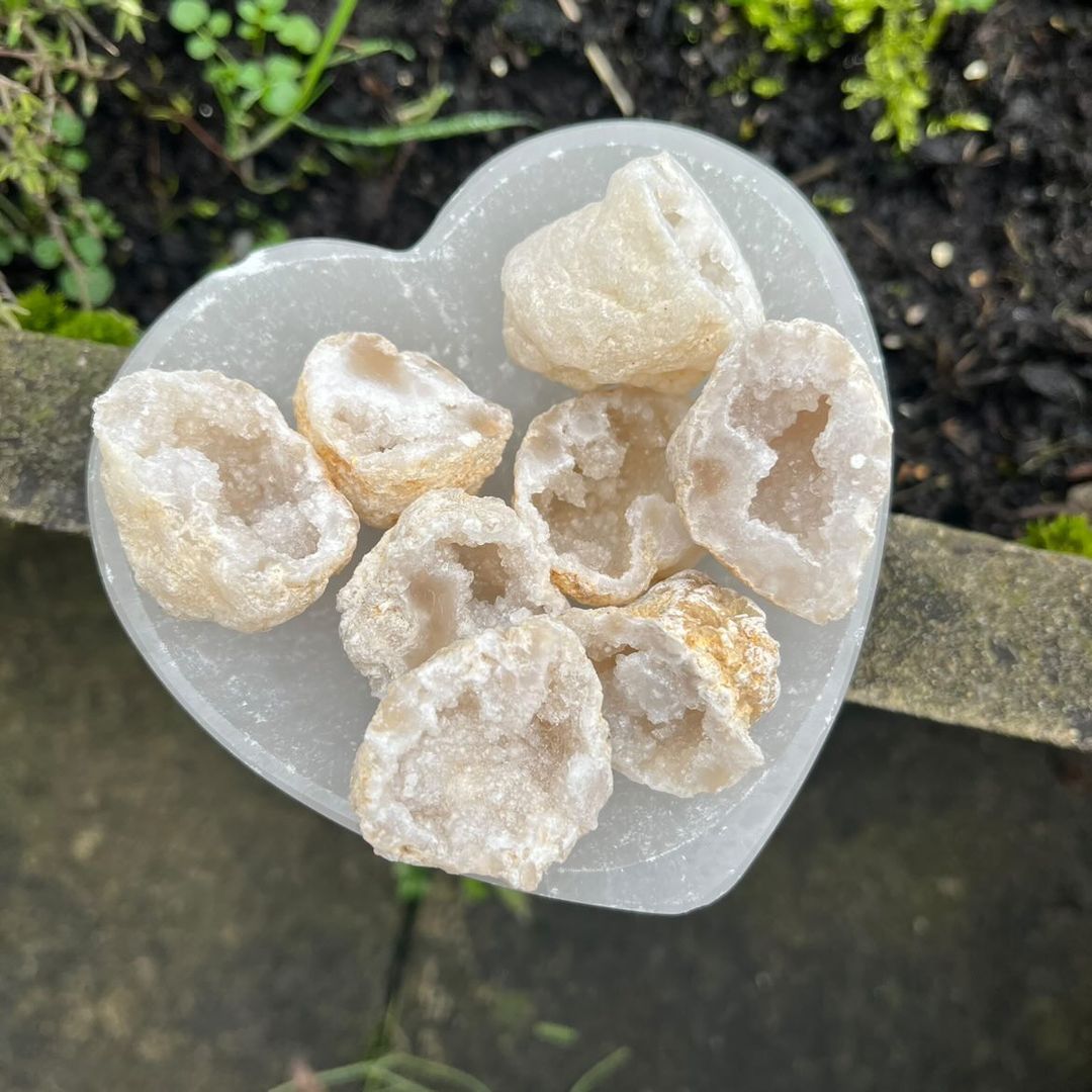 A collection of mini white quartz raw geodes gathered inside a translucent white Selenite heart bowl outdoors, showcasing their rough textured outer shells and glittering snowy interior crystal clusters.