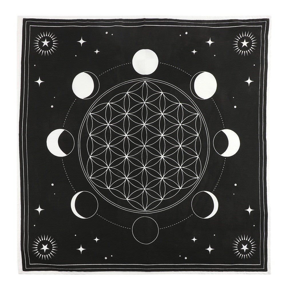 A top-down view of a square black cotton Moon Phase altar cloth laid flat against a clean white backdrop. The fabric features a central white Flower of Life geometric crystal grid, beautifully bordered by the changing cycles of the lunar moon phases, small stars, and sun graphics in the corners to support a mindful Third Eye and Crown chakra setup.