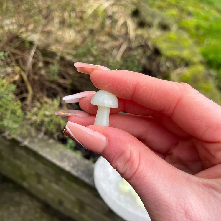 A close-up shot of a person gently holding a single pale green Jade mini mushroom crystal carving between their fingers outdoors. The natural light highlights its two-centimetre height, glassy smooth surface, and delicate internal veils against a blurred backdrop of dark soil and greenery to capture its portable everyday magic style.