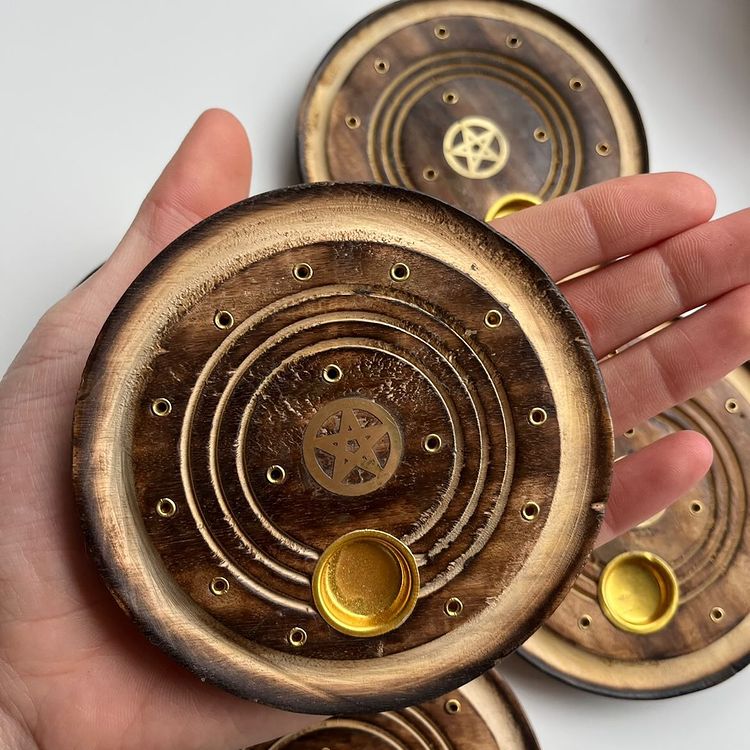 A close-up view of an open hand gently supporting a polished circular mango wood pentagram incense burner dish indoors. The perspective highlights the compact ten-centimetre kitchen or altar scale, smooth outer contours, and a bright gold-toned metal insert cup to emphasize its dual-purpose portable design for catching ash from favorite sticks and cones alike.