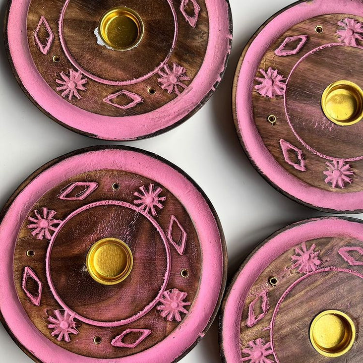 An overhead shot of four round mango wood incense burners featuring a bright pink painted rim, carved diamond and floral patterns, pre-drilled stick guide holes, and a central brass cup for burning incense cones safely.