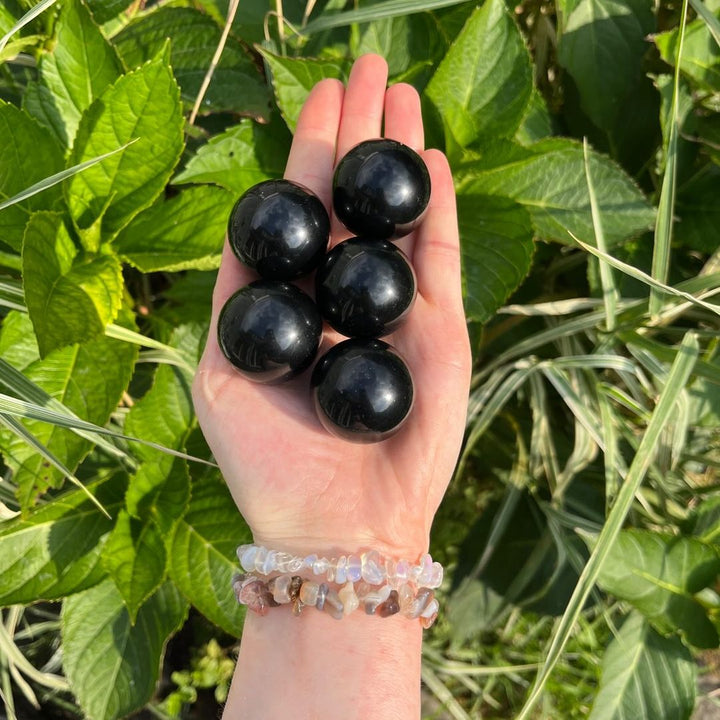 An open palm holding a collection of five small, glossy Black Obsidian crystal spheres outdoors, illustrating the compact 3cm to 4cm pocket-sized scale of these volcanic orbs against green garden leaves.