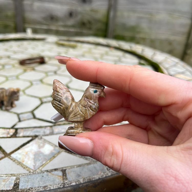A close-up view of a person gently holding a single, pocket-sized brown carved soapstone cockerel between their fingers outdoors. The shot highlights the miniature scale, detailed wing textures, and smooth tactile quality of the genuine stone bird against a softly blurred background of a rustic garden patio setup.
