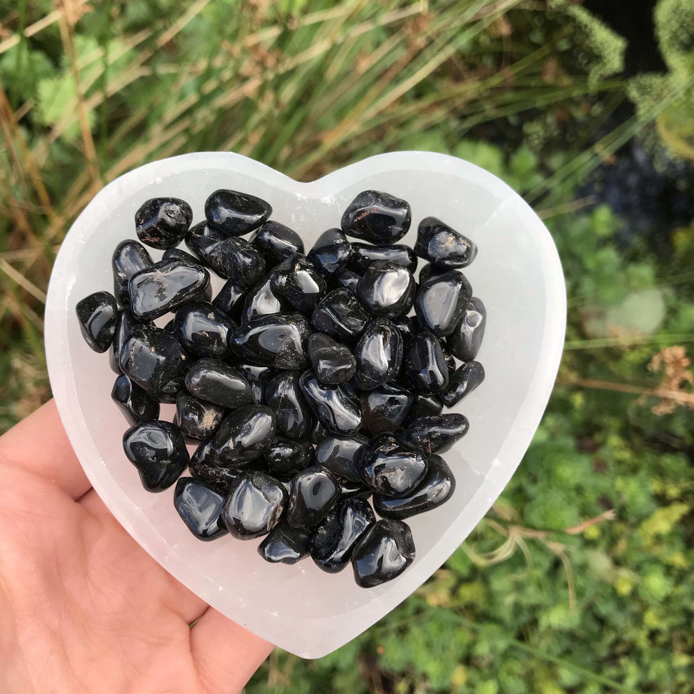 An outdoor shot showing a hand gently cradling a heart-shaped crystalline dish filled to the brim with tiny, sleek Black Onyx tumble stones. The background is a soft-focus texture of wild green garden grass, emphasizing the raw connection to nature and everyday lifestyle mindfulness.