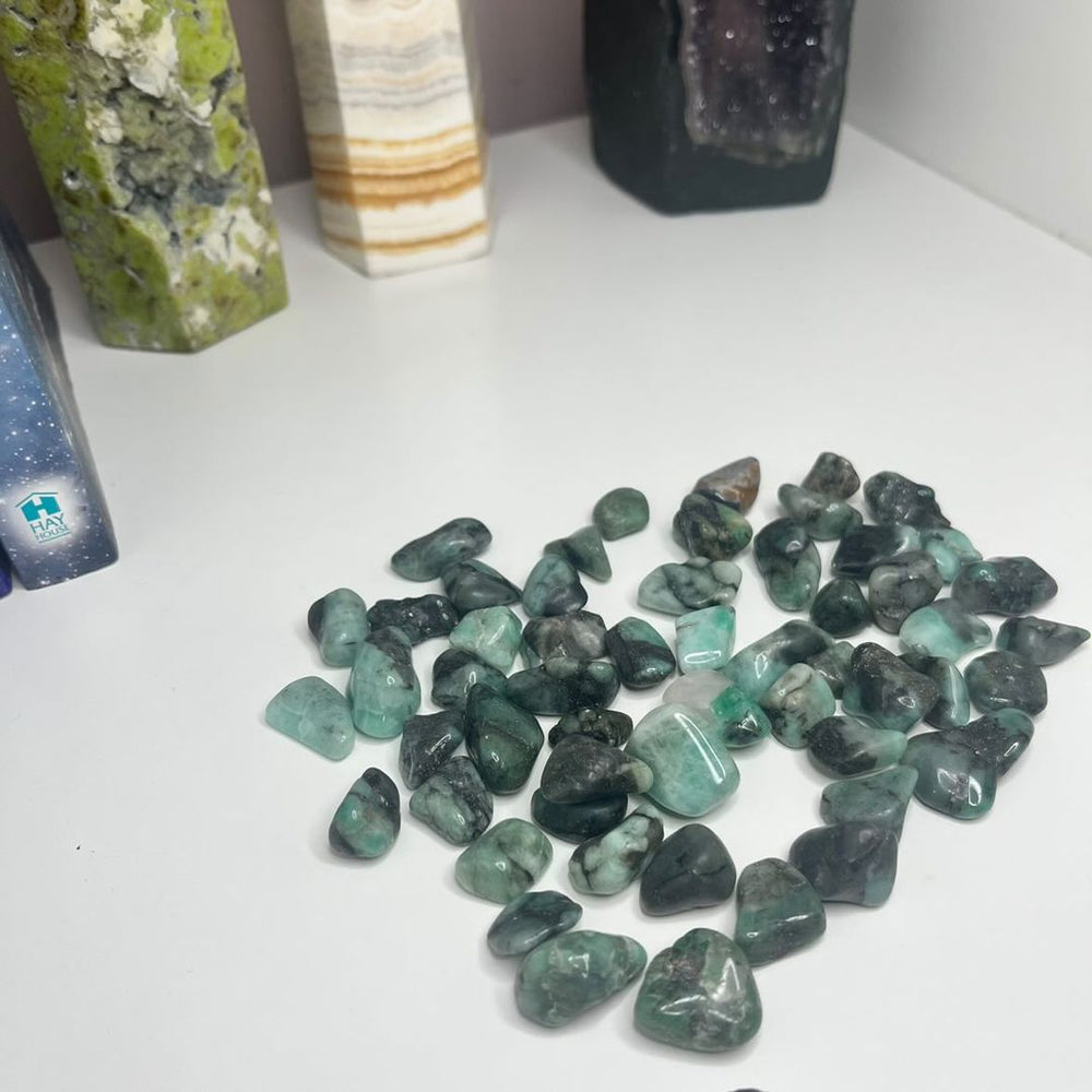 A high-angle view of a scattered pile of tiny deep-green polished emerald mini tumble stones on a clean white background, detailing the natural internal fractures and dark mineral birthmarks that promote emotional rejuvenation.