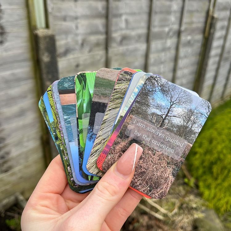 An outdoor close-up view of a hand with long, elegant nude nails holding a fan of Positive Affirmation cards. The crisp deck features high-quality photographic prints of nature scenes, trees, and changing seasons, displayed against a blurred background of a rustic wooden garden fence to welcome a comforting sense of mindfulness, emotional healing, and Crown chakra clarity.