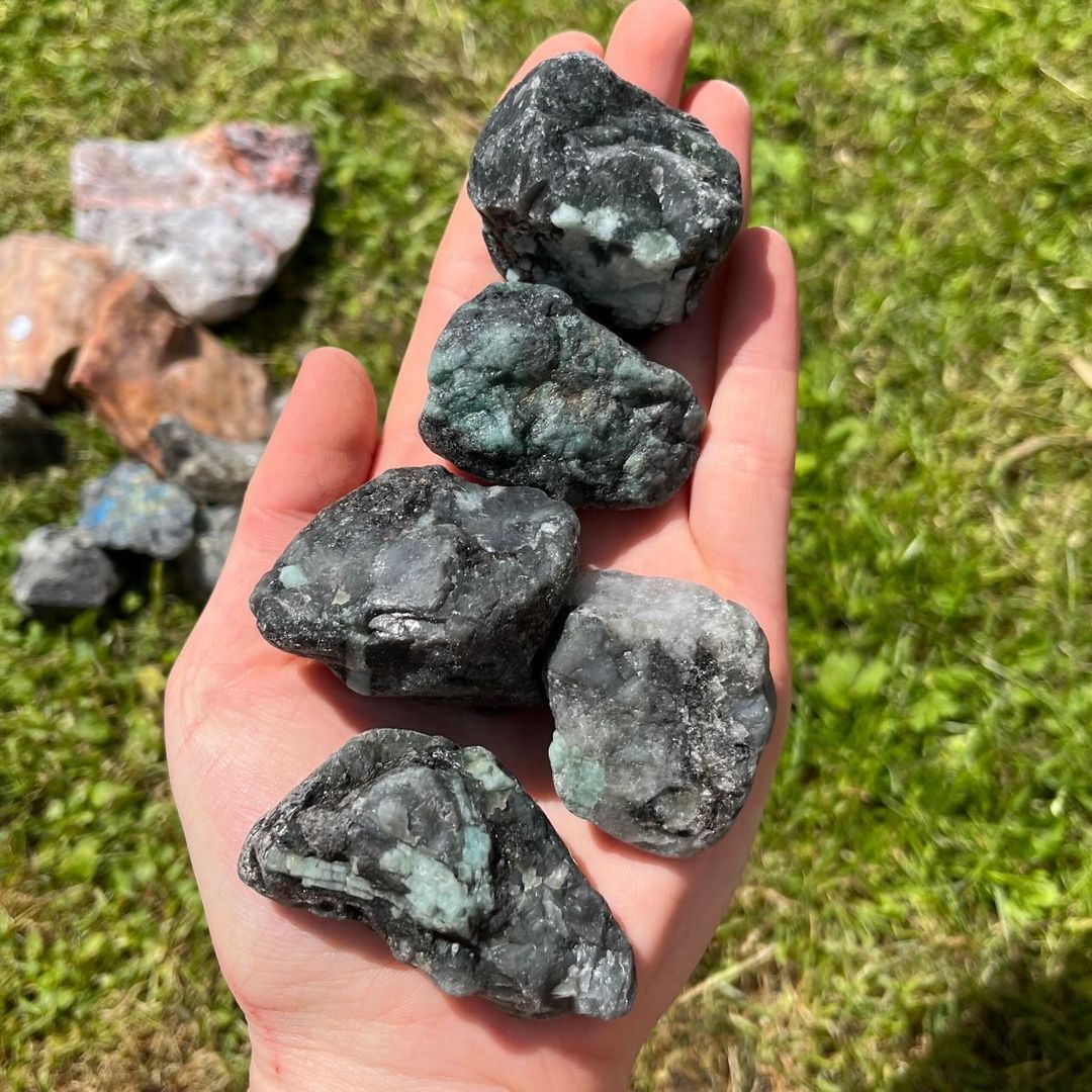 An open hand holding five unpolished raw emerald crystal specimens outdoors, displaying their rich green mineral inclusions, earthy black patterns, and rugged natural textures over sunny garden grass for Heart Chakra alignment.