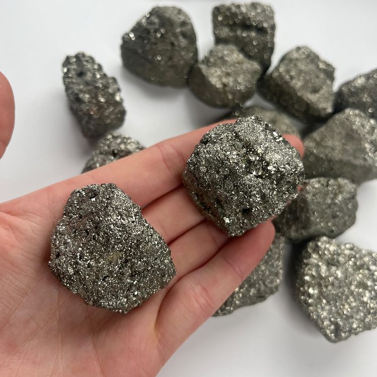 A hand holding two rough, glittering raw pyrite crystal chunks over a pile of fools gold specimens on a clean white background.