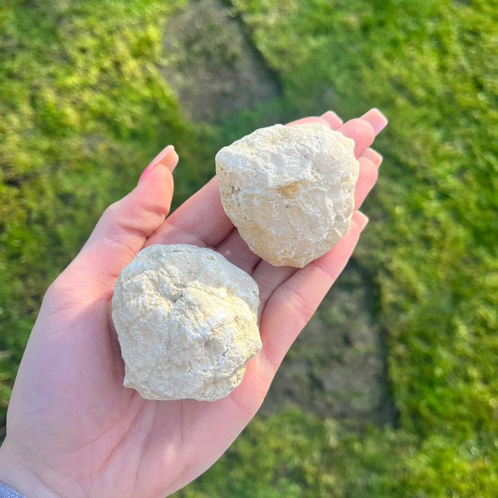 An open hand holding two large uncracked white quartz geodes under clear sunlight, highlighting the irregular geological shapes and earth-born features that provide a playful crystal experience for families.