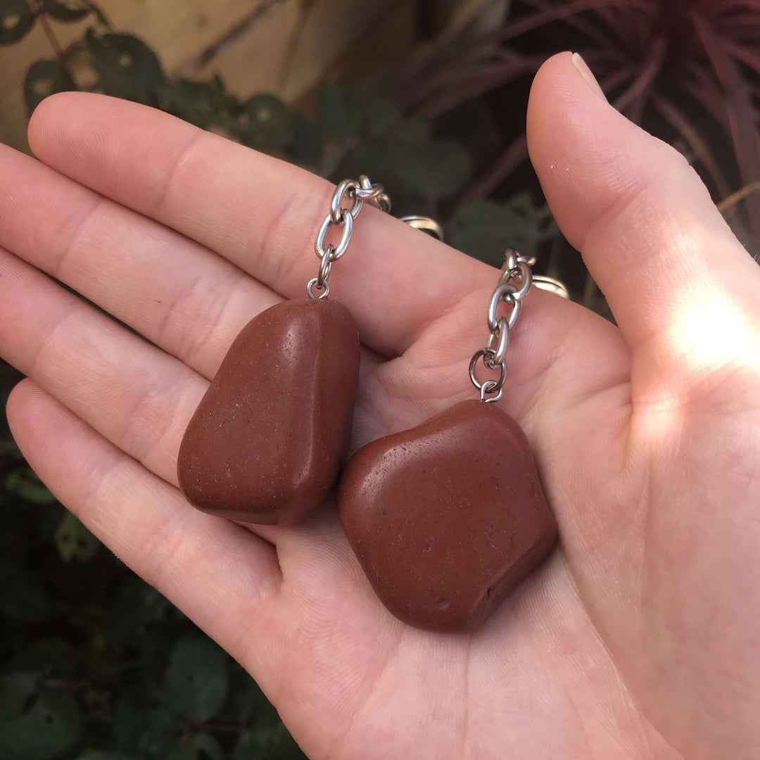 A close-up of polished, brick-red Red Jasper keyrings on an open palm, highlighting earthy textures and silver hardware for Root chakra grounding.