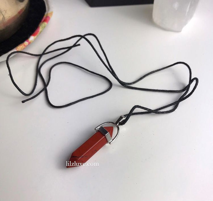A polished Red Jasper crystal point pendant in a silver metal bail setting, laid flat on a white desk with its long black cord spread out.
