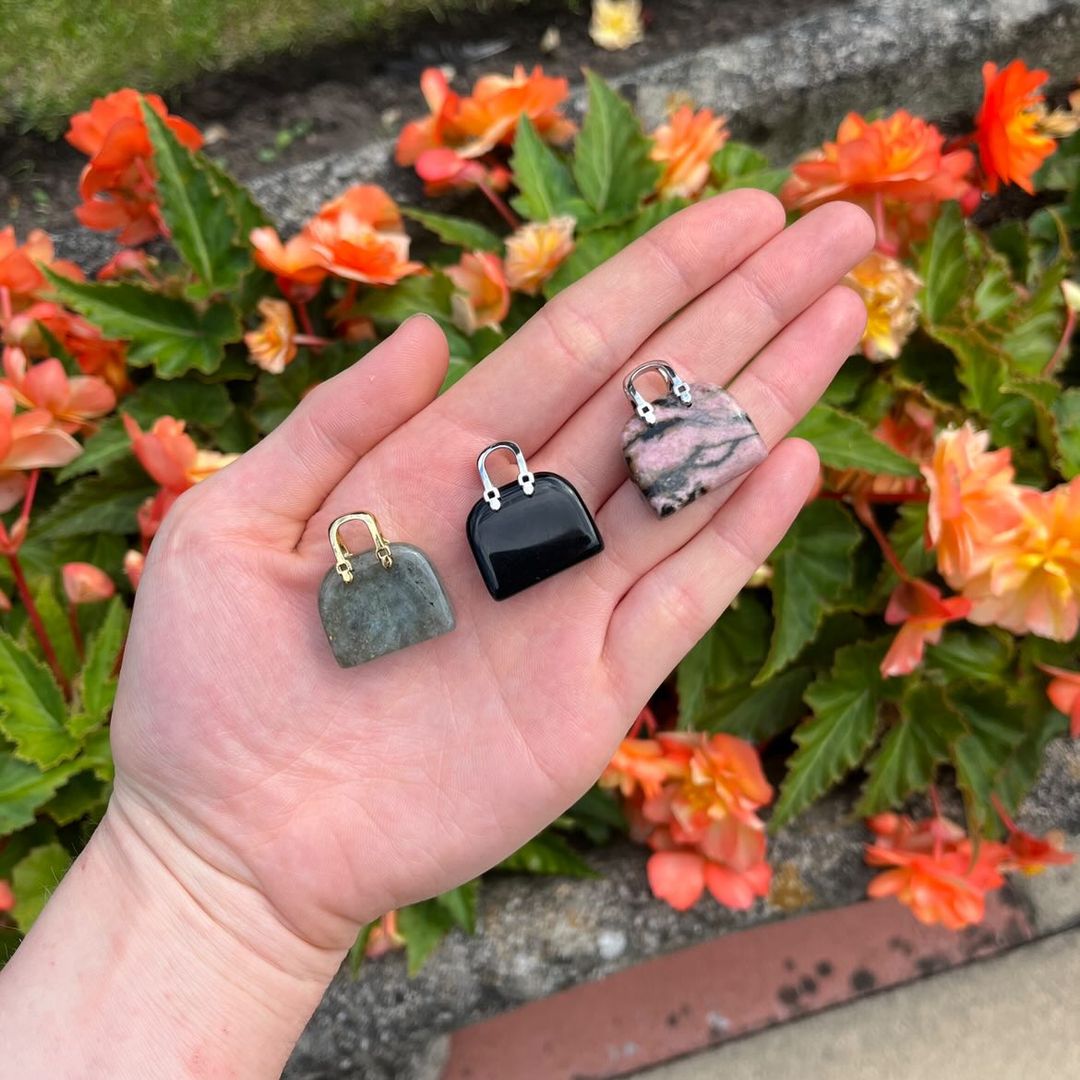 An open hand holding three tiny, polished crystal handbag carvings outdoors, showcasing a grey‑green flashes Labradorite piece, a jet‑black Obsidian purse, and a pink and black webbed Rhodonite token against bright orange garden flowers.